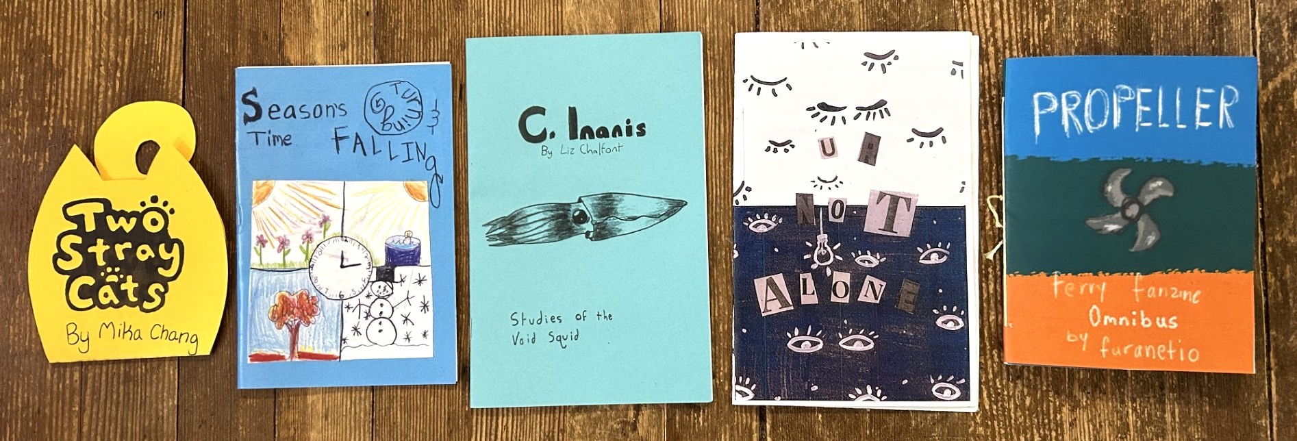 a photo of the 5 winning zines for the 11th annual wa state zine contest. two stray cats by mika change. Seasons Turning & Time Falling by Norah Armstrong. C. Inanis: Studies of the Void Squid by Liz Chalfont. Ur not Alone by Scarlette Kennard & Raquel Castillo. and Propeller: Ferry Fanzine (Omnibus.) by Furanetio.